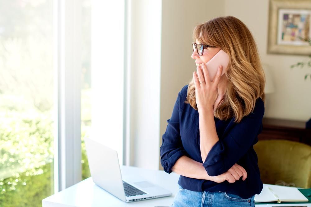 A Woman Is Talking On A Cell Phone While Standing In Front Of Laptop — Howells & Co Legal & Conveyancing In Gosford, NSW