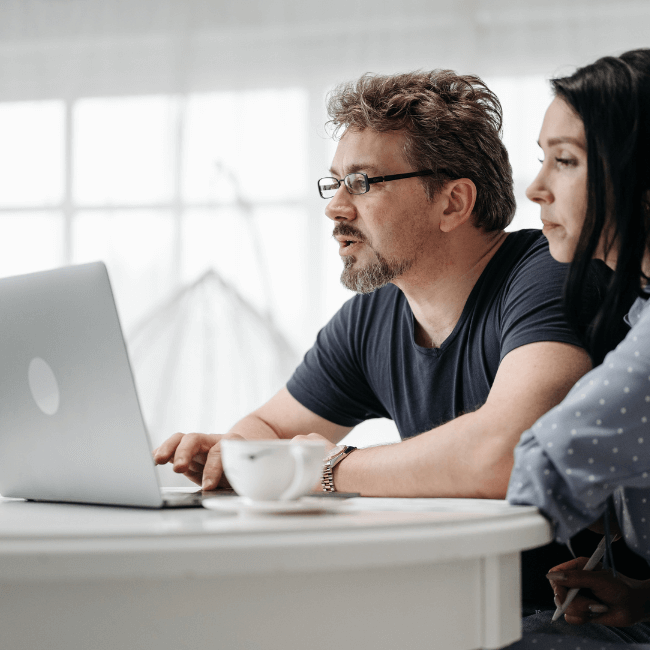 A Man And A Woman Are Sitting At A Table Looking At A Laptop — Howells & Co Legal & Conveyancing In Cessnock, NSW