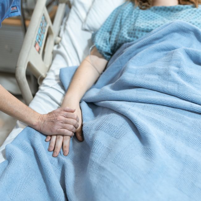 A Woman Is Laying In A Hospital Bed Holding Another Woman's Hand — Howells & Co Legal & Conveyancing In Cessnock, NSW