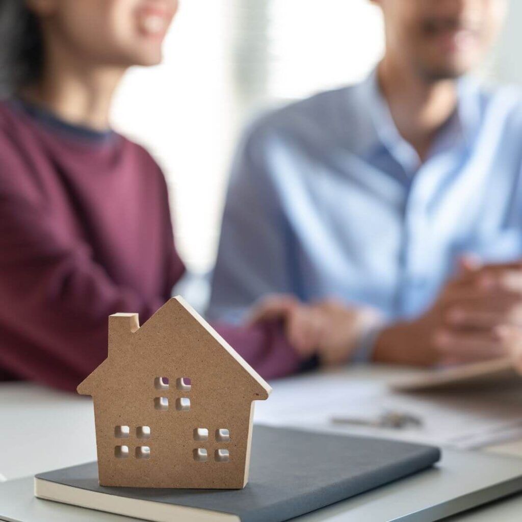 A Man And A Woman Are Sitting At A Table With A Model House On Top Of A Notebook — Howells & Co Legal & Conveyancing In Cessnock, NSW