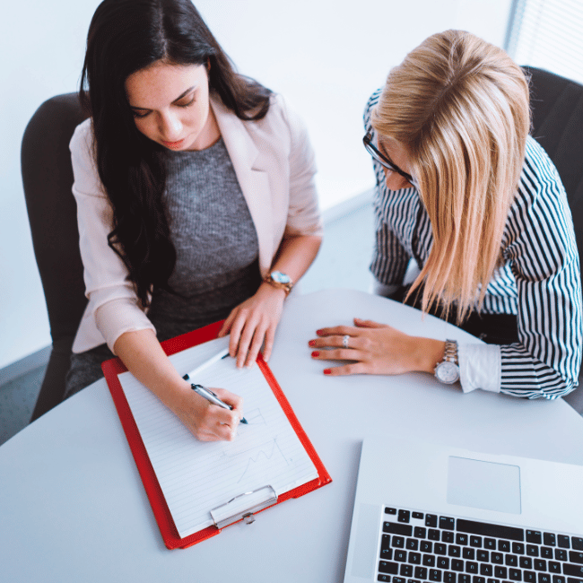 Two Women Are Sitting At A Table With A Clipboard And A Laptop — Howells & Co Legal & Conveyancing In Cessnock, NSW