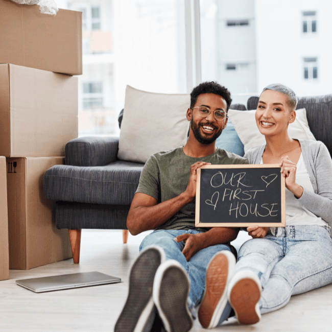 A Man And A Woman Are Sitting On The Floor Holding A Sign That Says Our First House — Howells & Co Legal & Conveyancing In Cessnock, NSW