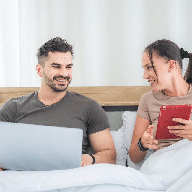 A Man And A Woman Are Sitting In Bed Looking At A Laptop And A Tablet — Howells & Co Legal & Conveyancing In Cessnock, NSW