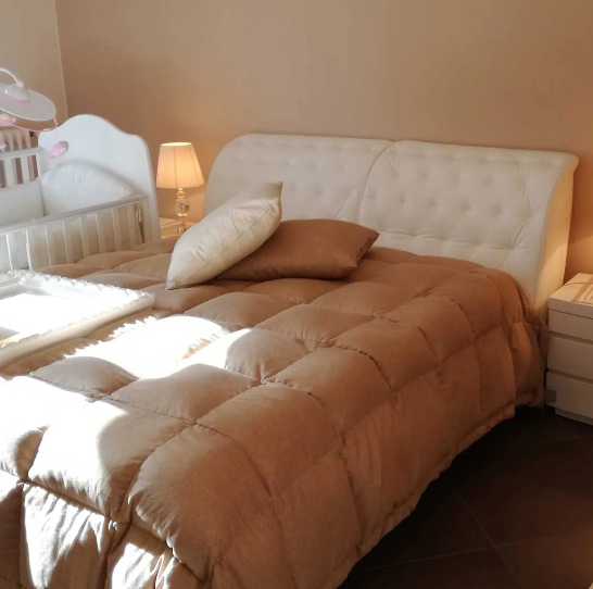 Accogliente camera da letto beige con letto trapuntato, piumone color cuoio, cuscini e lampada da comodino, illuminata da una calda luce solare.