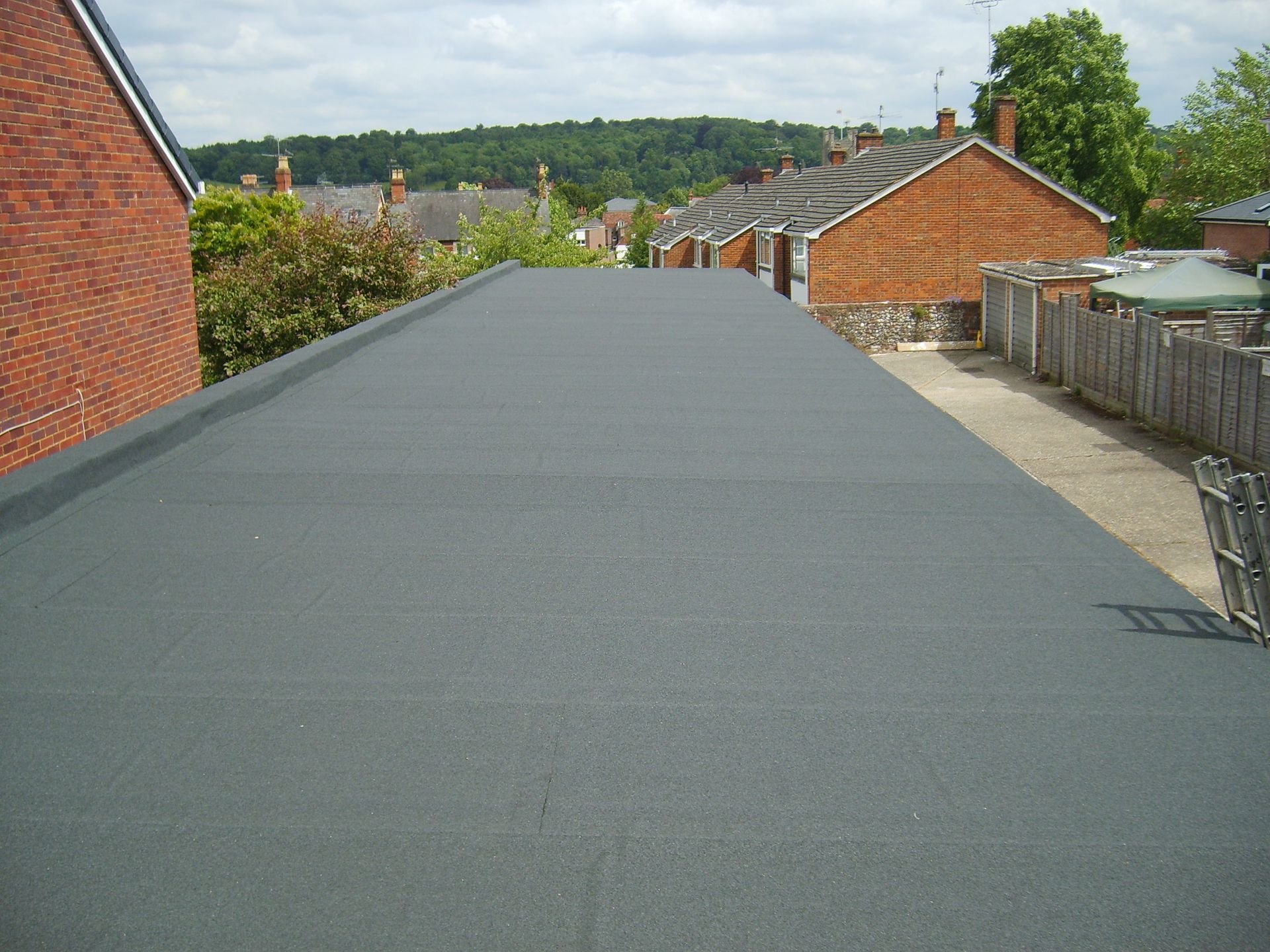 Flat, dark grey roof with houses in the background. Clear day with a treeline visible in the distance.