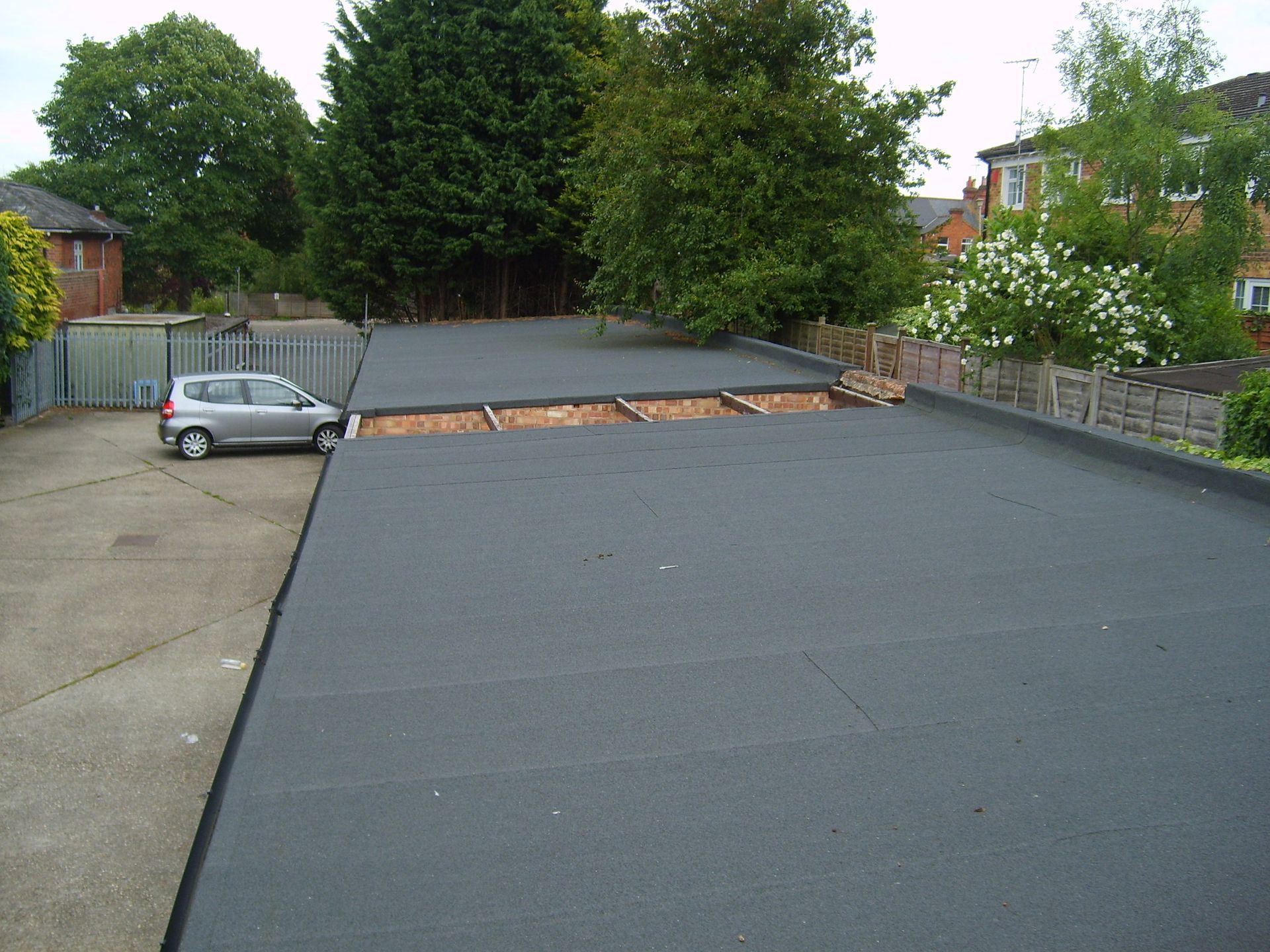 A gray car parked on a paved area next to a flat, dark-colored roof. Trees and houses are visible in the background.