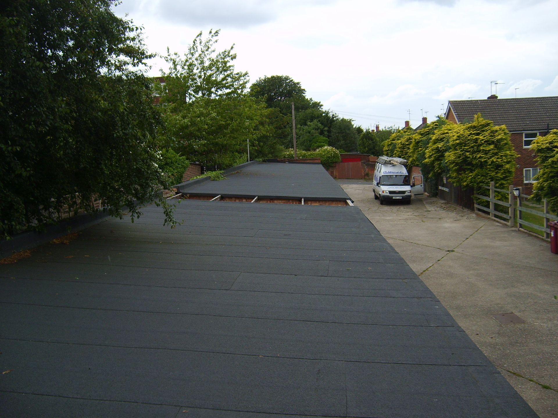 A long, black, flat roof with a utility van parked on a driveway next to a row of houses. Trees line the background.