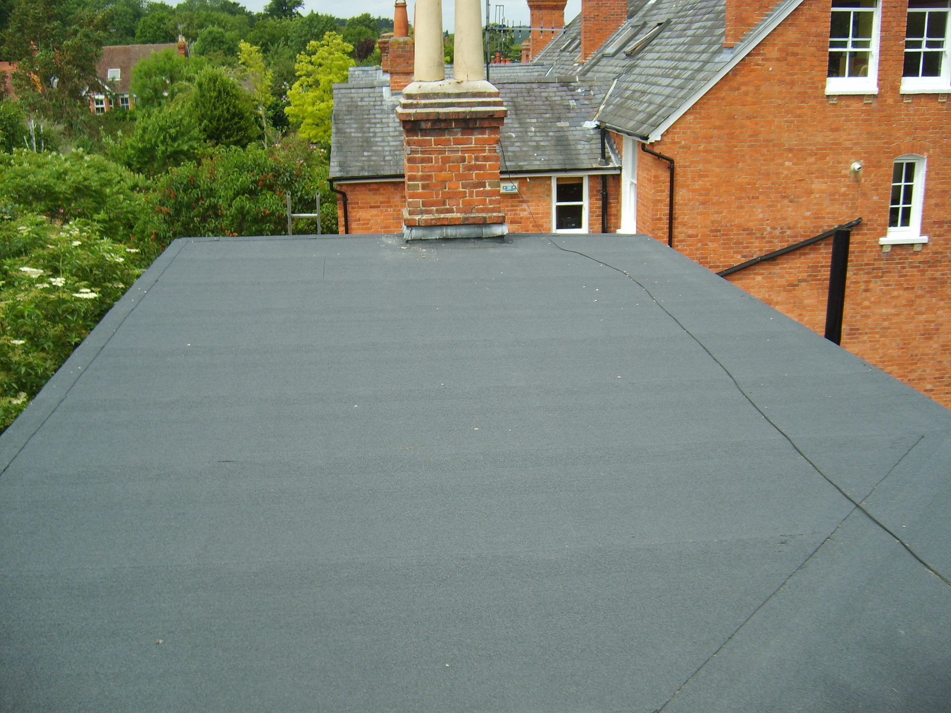 Black flat roof with a brick chimney against a backdrop of trees and a red brick building.