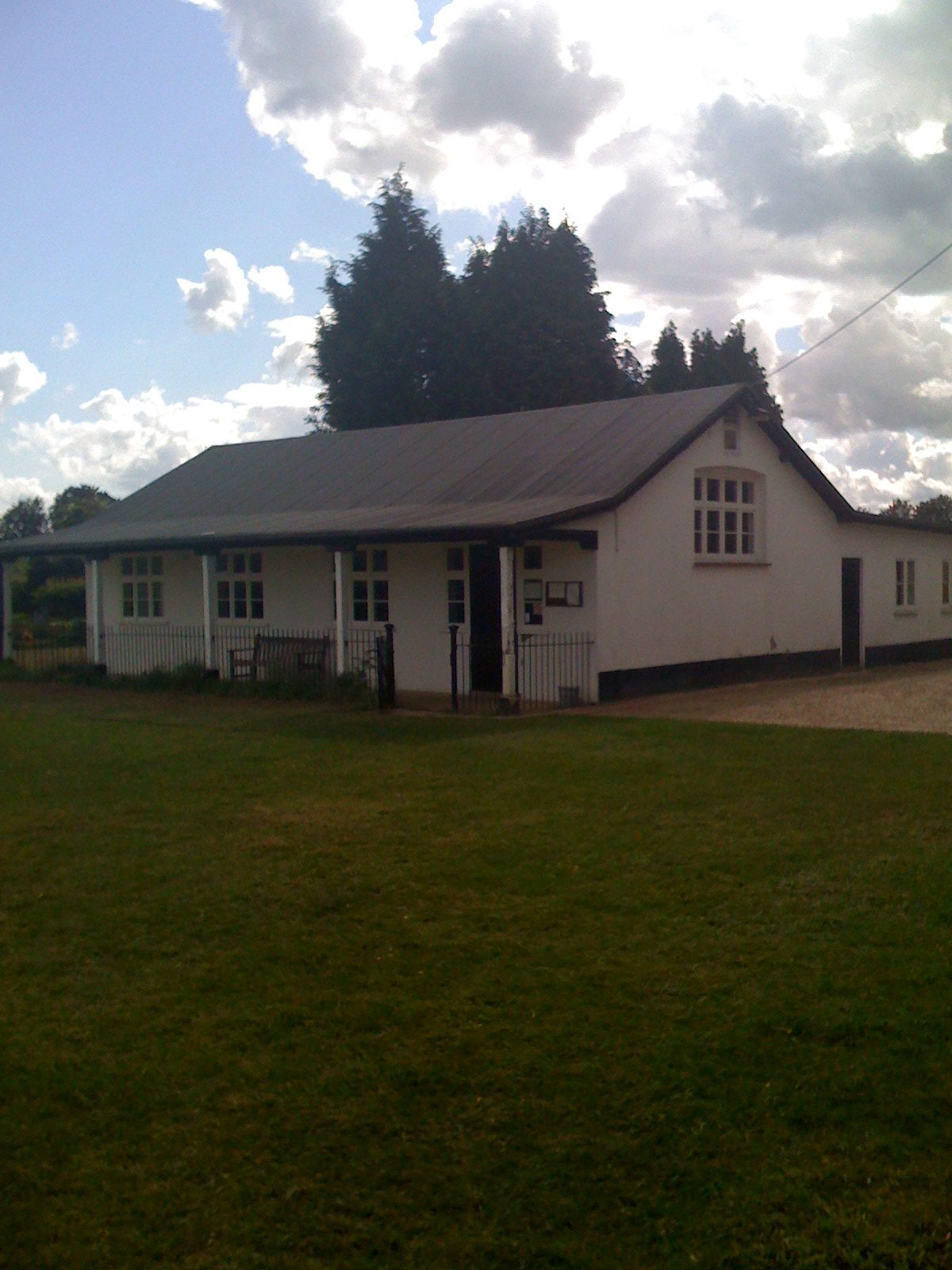White building with a dark roof and multiple windows on a green field, under a cloudy sky.