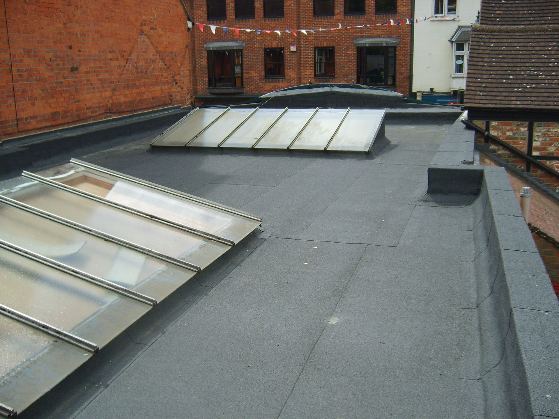 A flat roof with two skylights, brick wall, and a glimpse of surrounding buildings. The roof is dark grey.