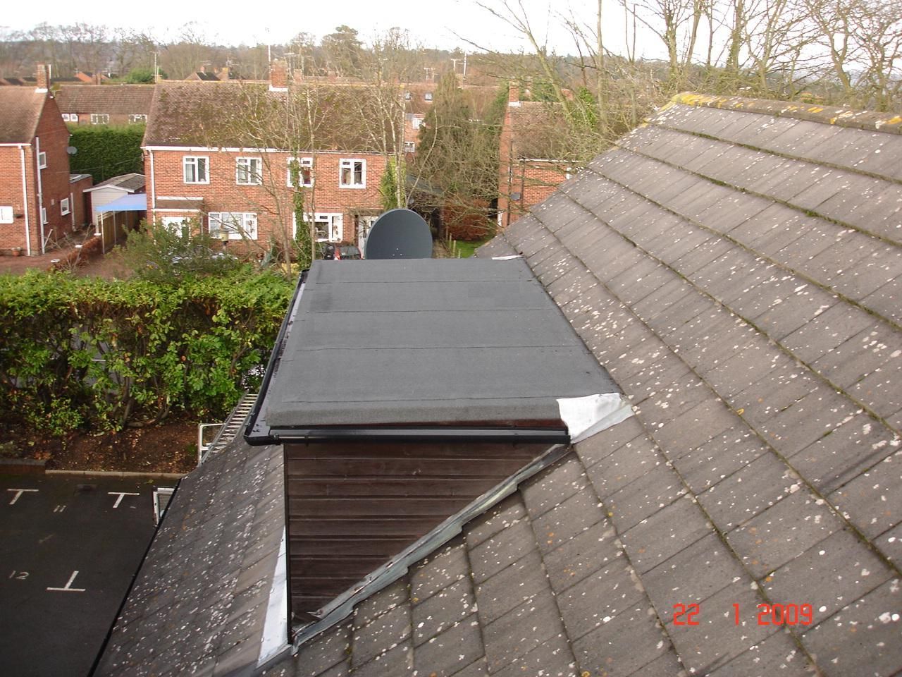 A rooftop view showing a flat black roof section next to a sloped, tiled roof. Houses and trees are in the background.