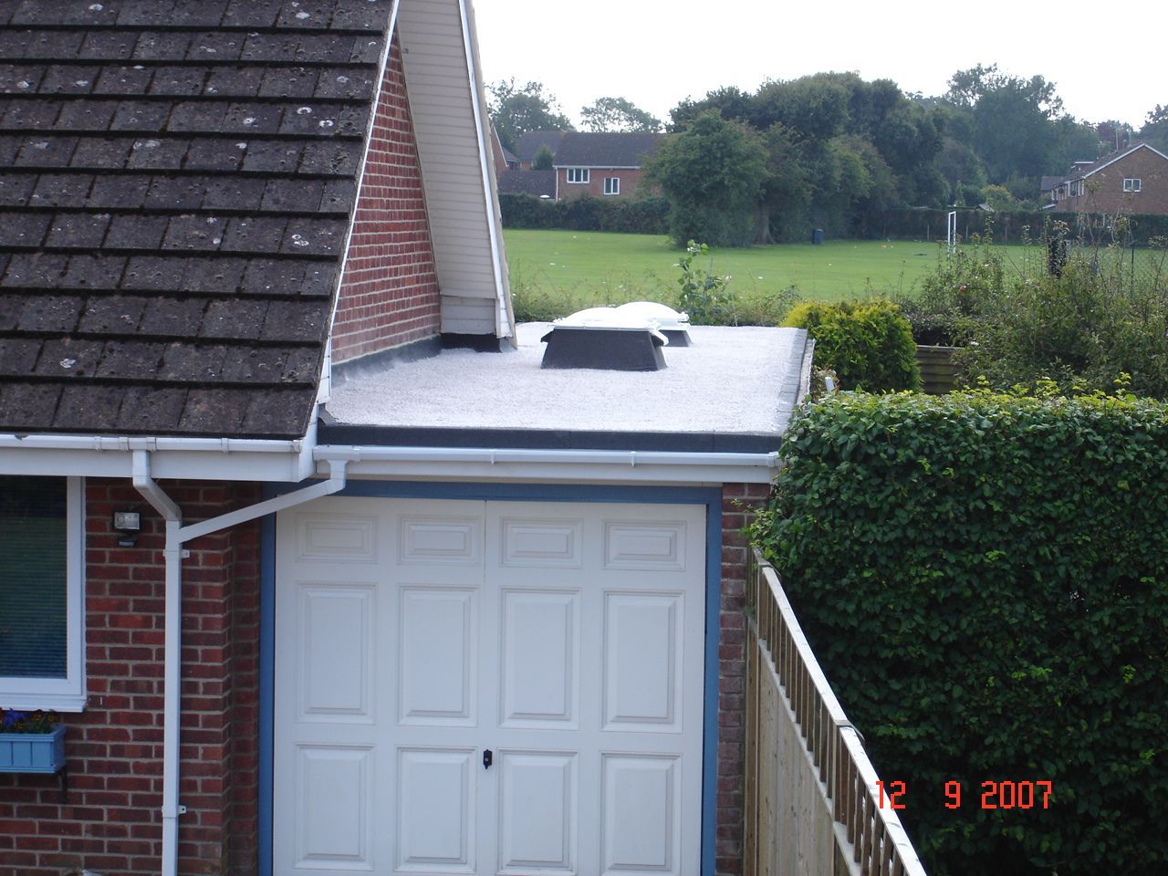 Flat roof of a garage with two skylights, white garage door, and a brick house next to a grassy yard.