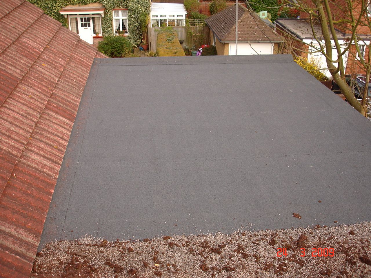 Flat, black roof surface next to a reddish-brown tiled roof. Houses and trees visible in the background.