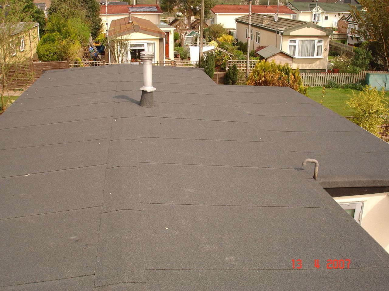 Flat, dark roof with a chimney, overlooking a residential area with houses and greenery.