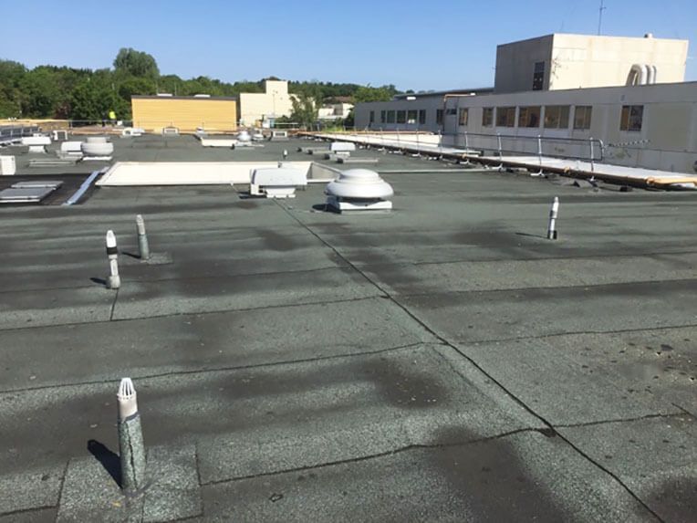 View of a flat, dark green commercial roof with vents, pipes, and a building in the background on a sunny day.