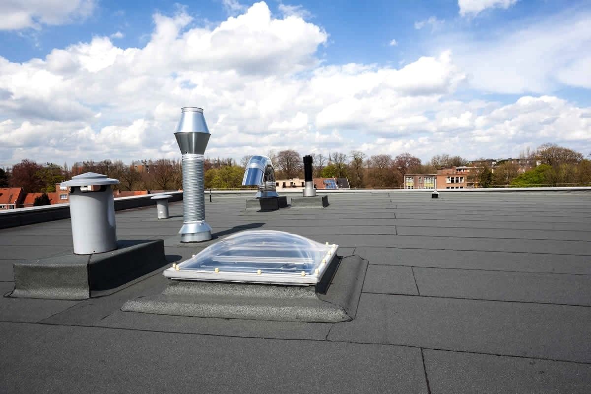 Flat, black commercial roof with vents, a skylight, and a blue sky. Trees and buildings are in the background.