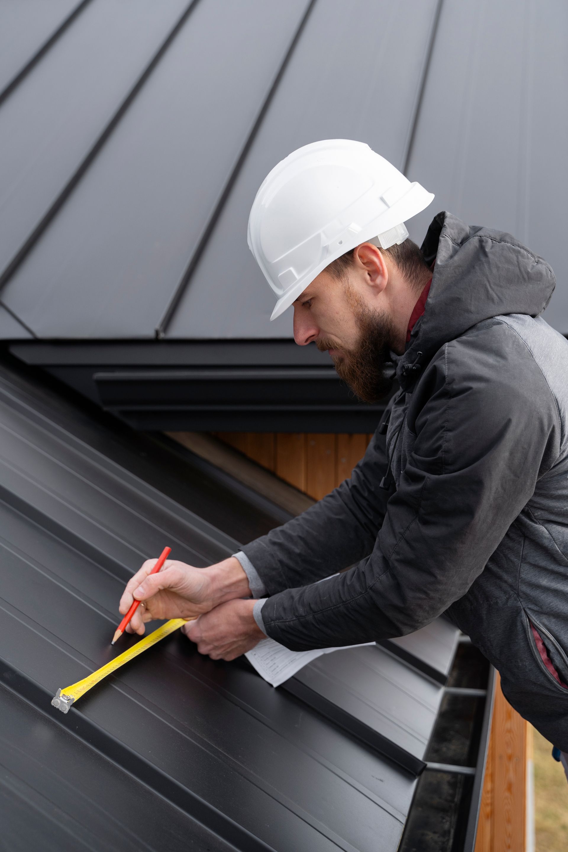Worker in a white hard hat measures a black metal roof with a tape measure.