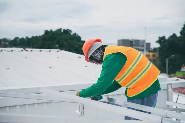 Worker in orange safety vest and hard hat inspecting a rooftop under cloudy skies