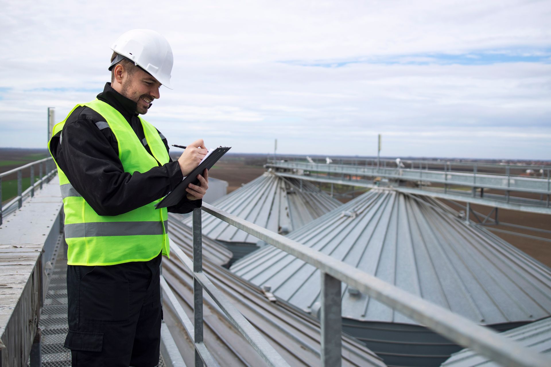 A man in a hard hat and vest inspects a metal silo, taking notes on a clipboard, smiling, and standing outdoors.