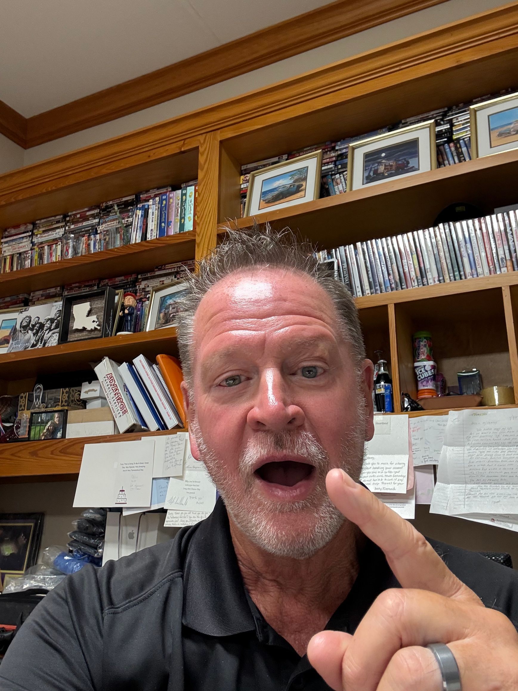 Man with open mouth, pointing finger, in front of a bookshelf filled with items.