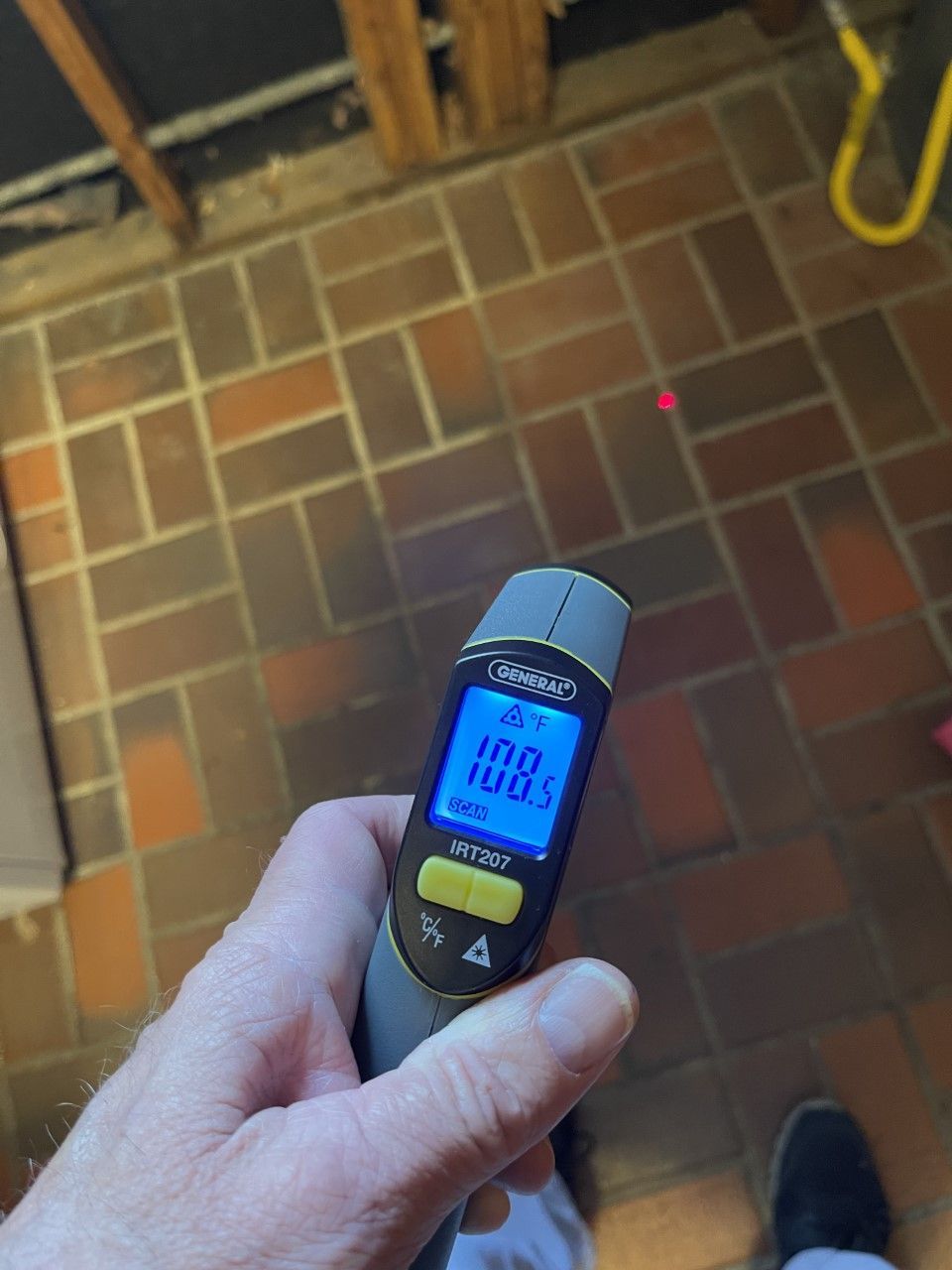 Hand holding a temperature gun displaying 108.1 degrees on brick floor. Red laser dot visible.