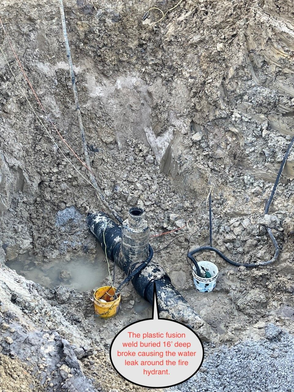 Worker in a deep muddy trench repairing a black pipe with tools and buckets nearby.
