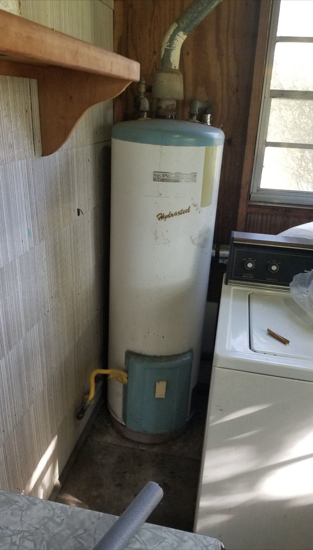 A tall, white and blue water heater inside a room, next to a washing machine.