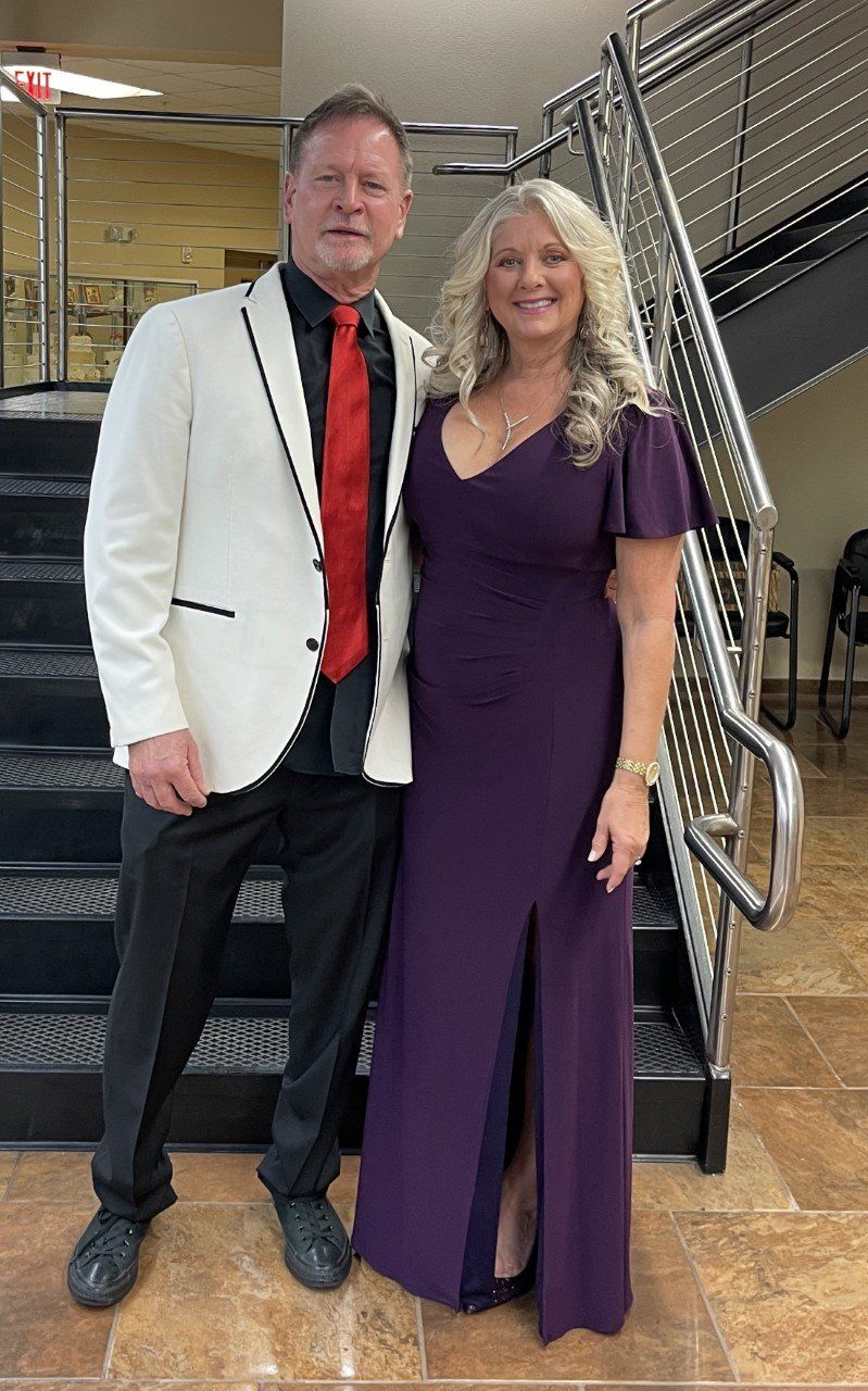 Two adults posing on a staircase, wearing formal attire; one in a white blazer, the other in a purple dress.