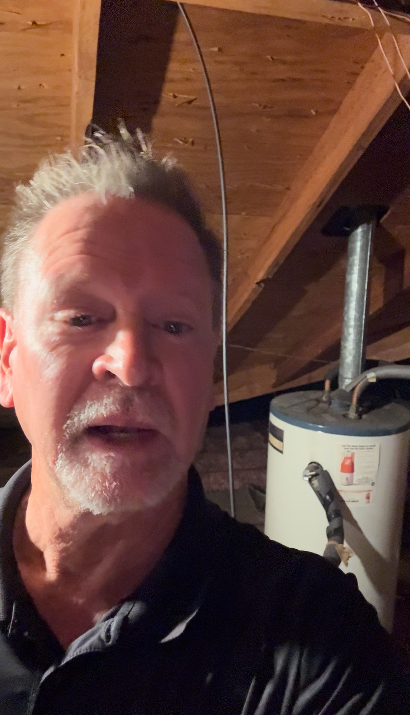 Selfie of a man in a black shirt in a wood-paneled attic with a water heater behind him