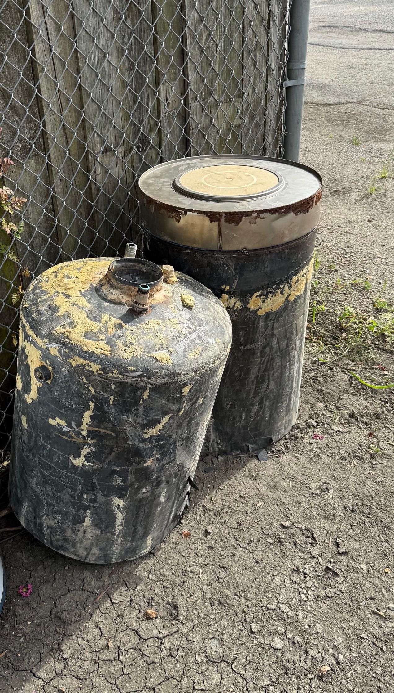 Two weathered black barrels beside a chain-link fence on gravel.