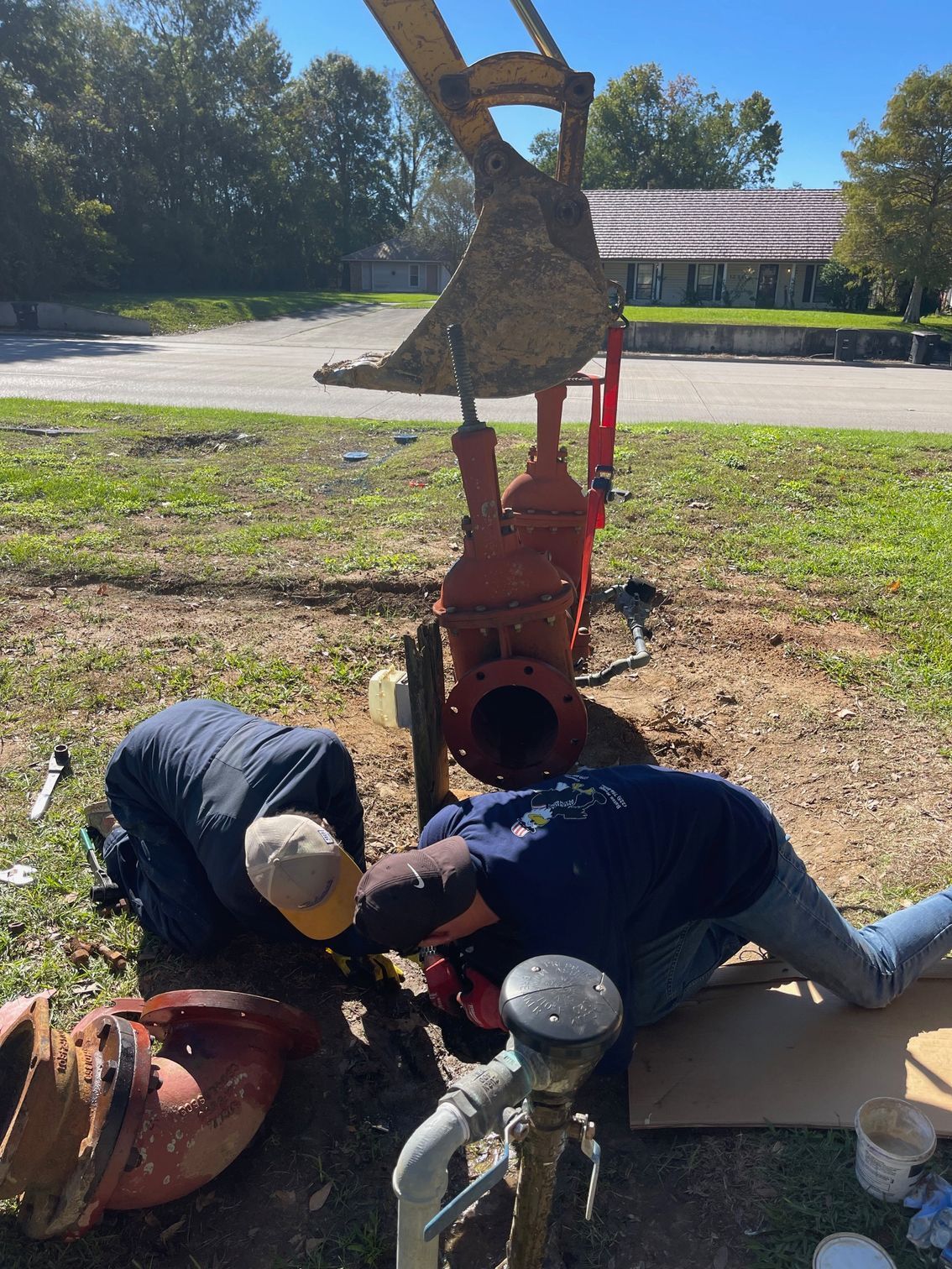 Workers crouch beside a red fire hydrant and pipe on a grassy roadside area.