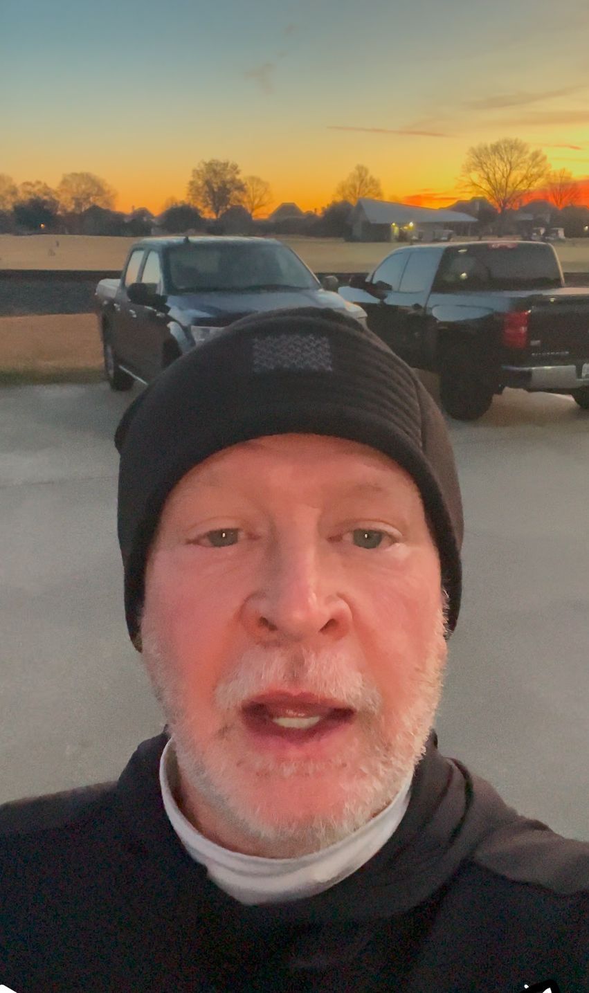 Close-up selfie of a man in a black beanie outdoors at sunset, with parked trucks behind him.