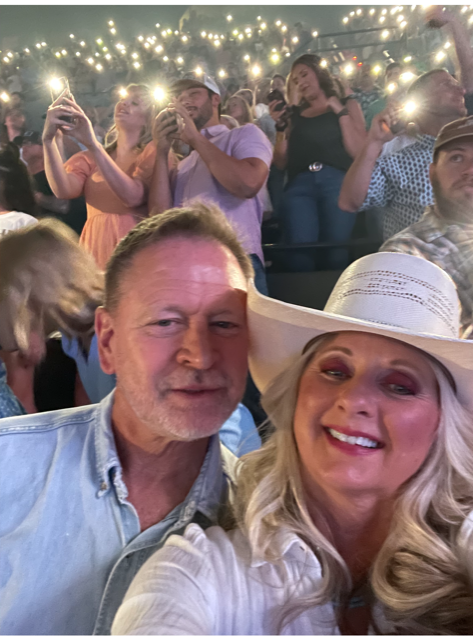 Smiling couple taking a selfie at a concert. Blonde woman in a white hat, man in blue shirt, crowd with lights.