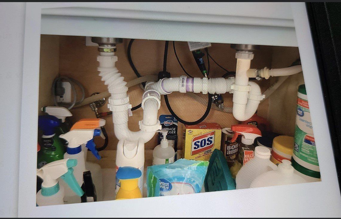 Under-sink cabinet with white plumbing pipes and cleaning supplies packed below the sink.