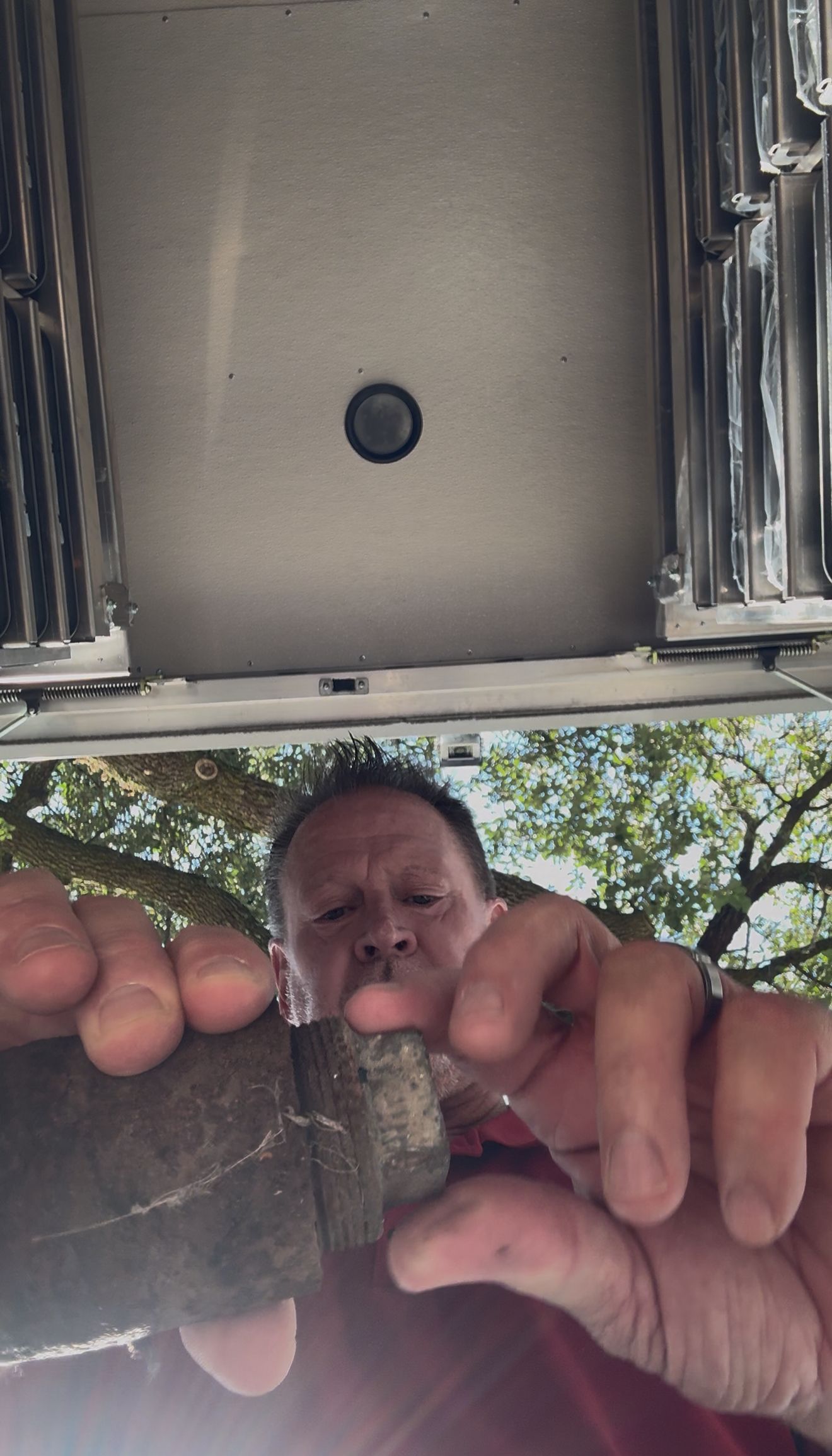 Close-up of hands holding a small object, with a man visible below beneath a metal structure outdoors