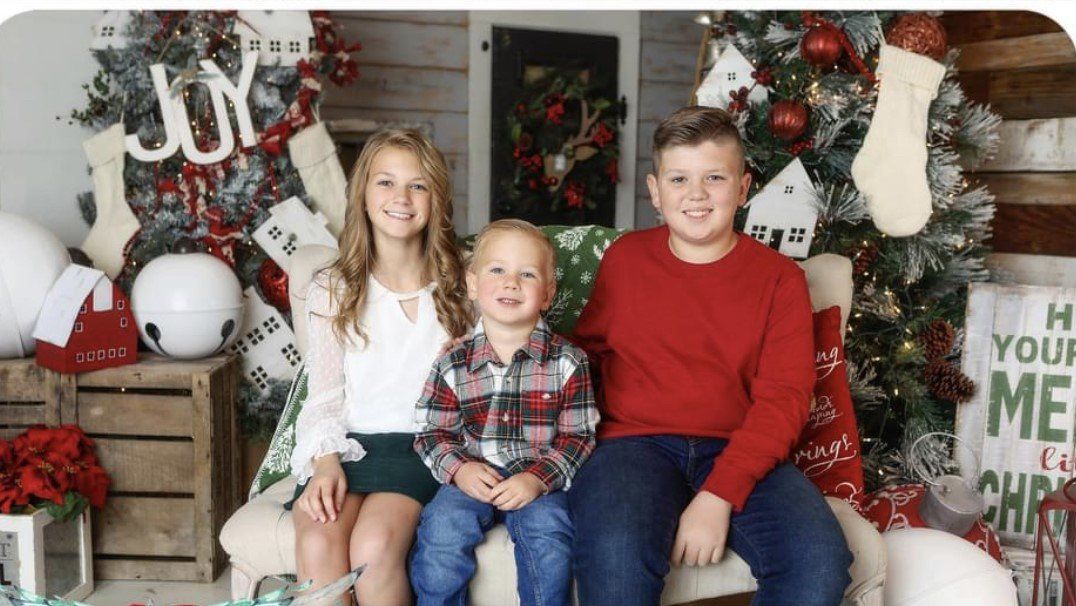 Three children smiling in front of a decorated Christmas tree. Girl in green skirt, boy in red sweater, other boy in plaid shirt.
