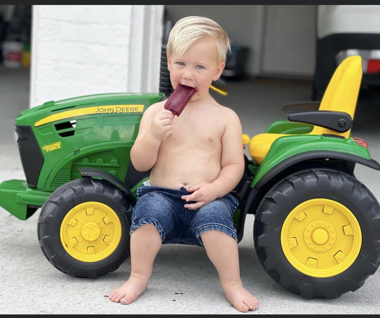 Blond toddler eating popsicle, shirtless, on a toy John Deere tractor, outdoors.
