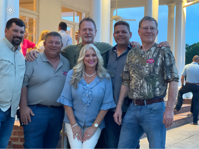 Group of six adults posing together outdoors at a porch entrance, smiling and dressed casually.