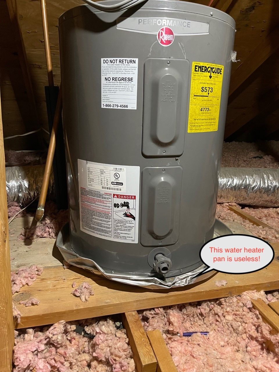 Gray water heater in a basement, with pink insulation scattered on the floor and warning labels on the tank.