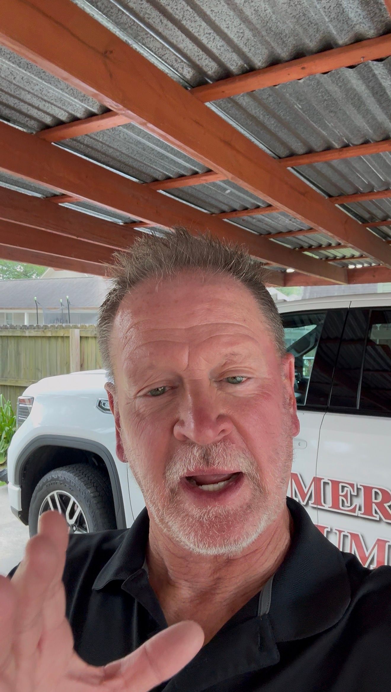 Man in black shirt speaking under a metal carport beside a white pickup truck