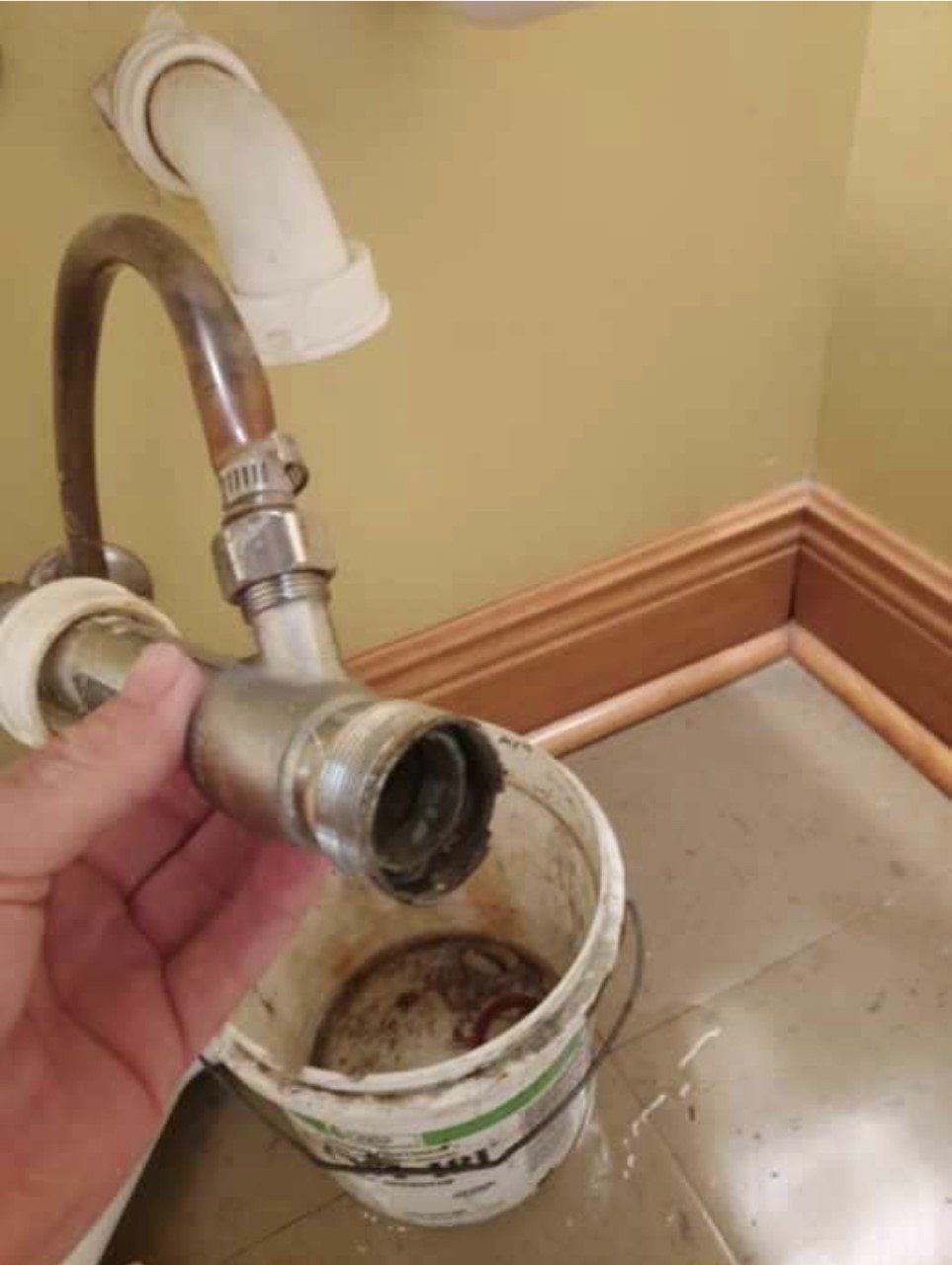 A hand holding a disassembled sink drain pipe over a bucket, indoors.