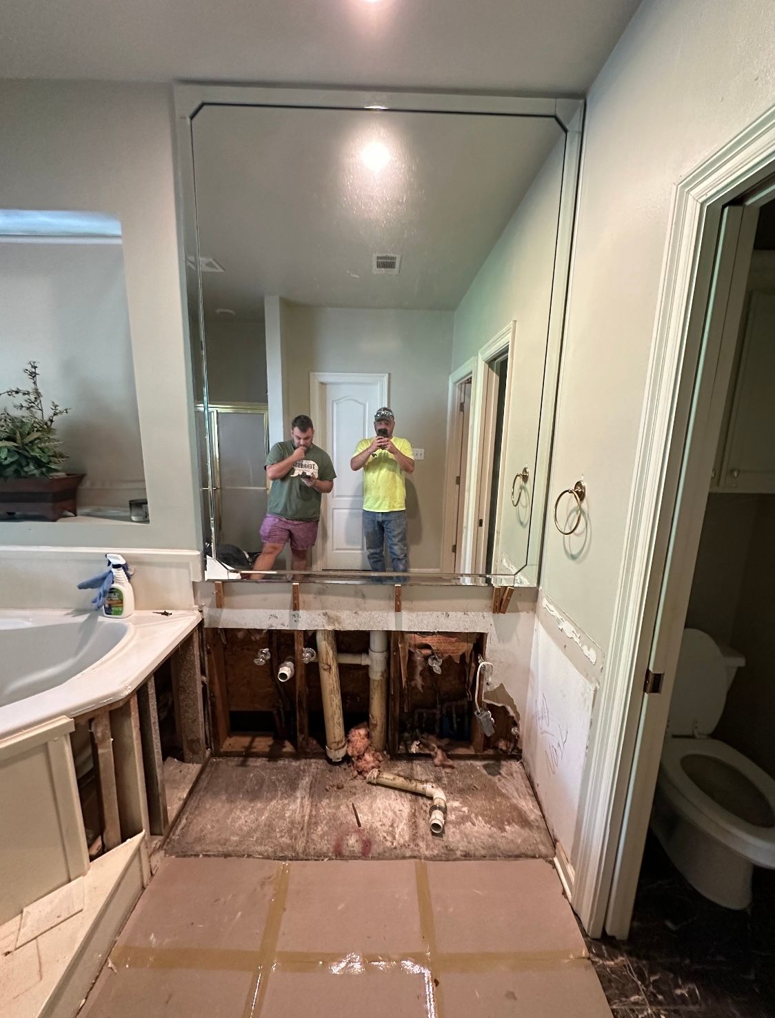 Bathroom renovation in progress. Exposed plumbing under a large mirror, two people taking pictures, and a toilet.