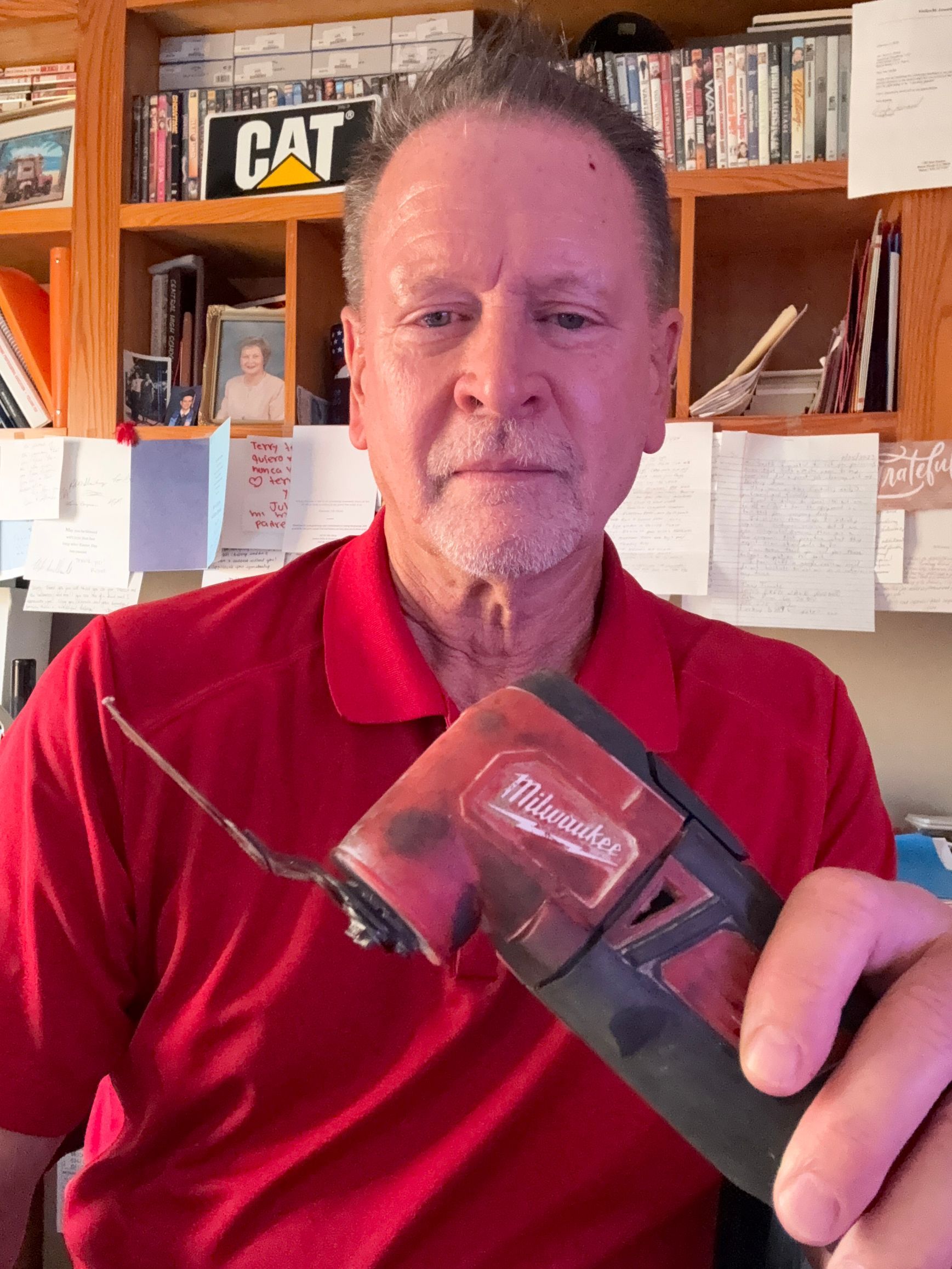 Man in red shirt holding a black device toward the camera, standing in a home office with shelves behind him