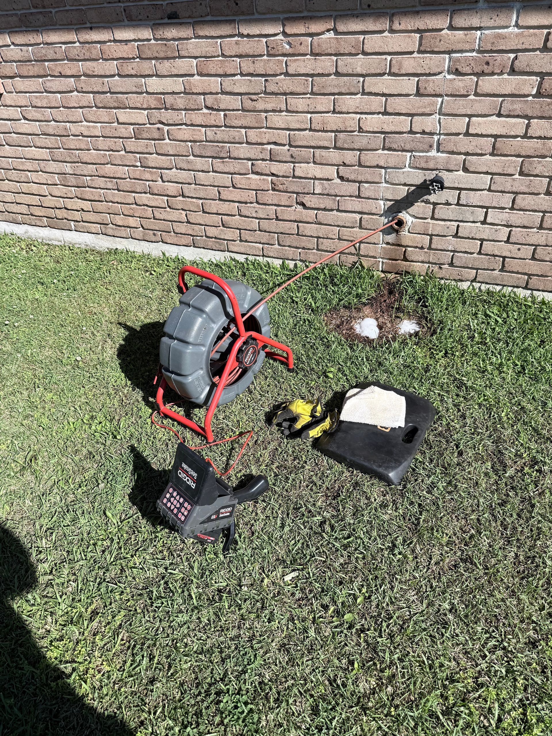 Backpack leaf blower, gloves, and helmet on grass beside a brick wall and faucet