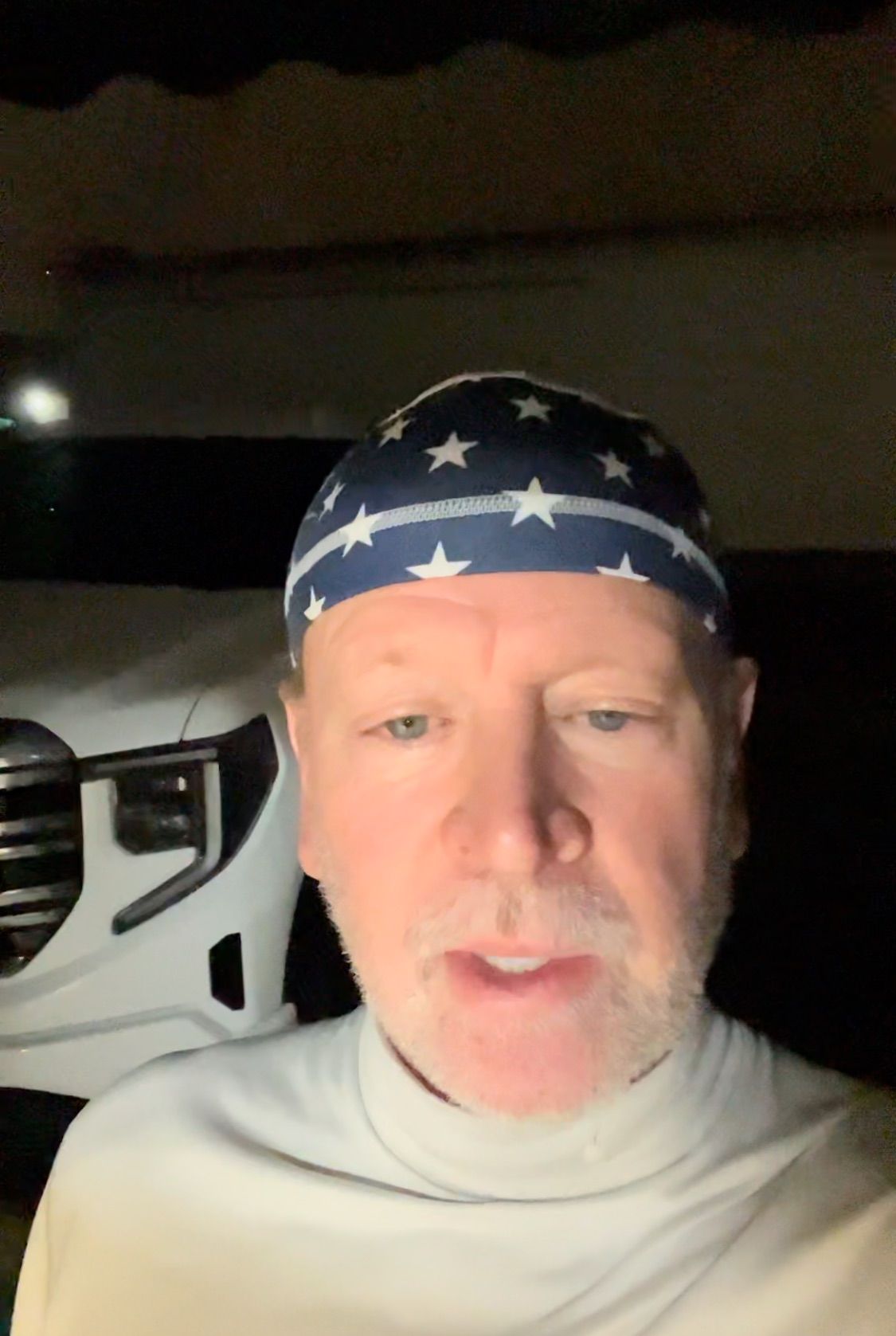 Man in a blue and white star bandana speaks outside a white truck at night.