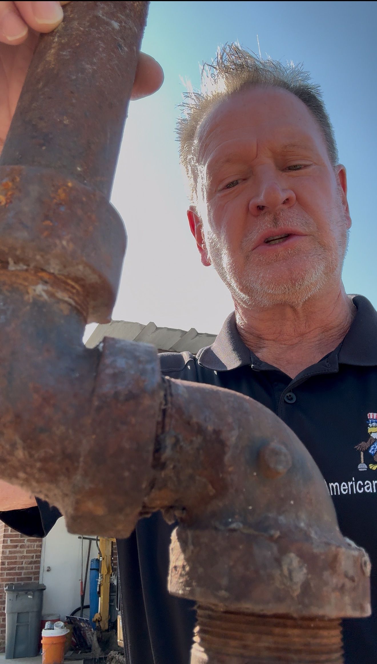 Man holding a rusty metal pipe elbow outdoors, talking to the camera.