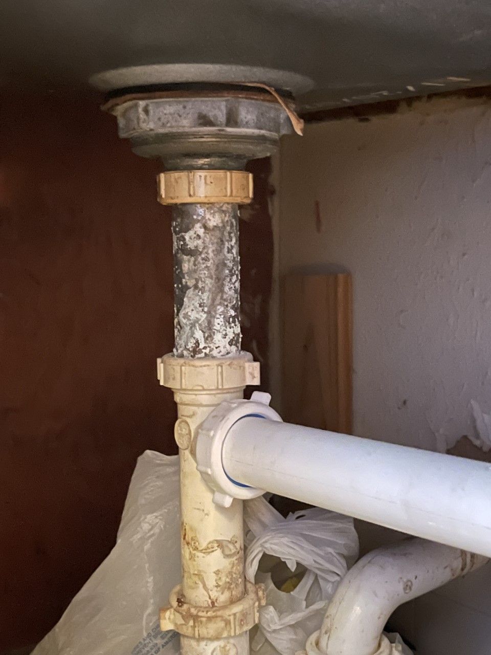 Under-sink plumbing with a corroded vertical pipe connected to a white drain pipe and fittings