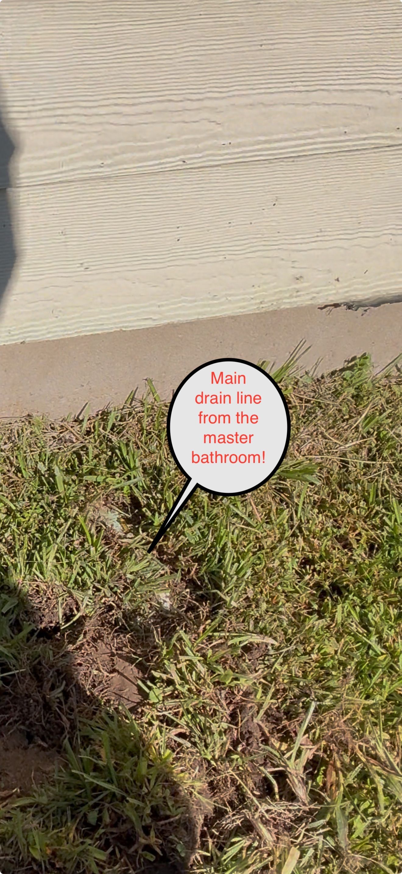 Small round sign with red text staked in a patch of grass beside a beige wall.