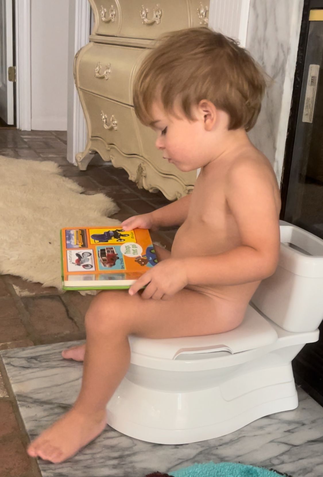 Child sitting on a small white potty, reading a colorful book in a bathroom.