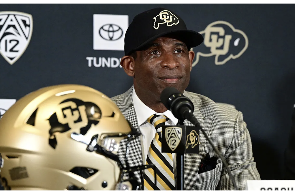 Deion Sanders in a black hat and suit at a press conference. A gold helmet sits to his left.